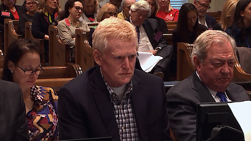Three people sit at a courtroom table, looking serious. They are surrounded by other people seated in wooden pews, suggesting a legal proceeding in progress. The atmosphere is formal and attentive.