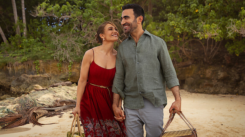 A happy couple holding hands and carrying beach bags, walking on a sandy beach with trees and rocks in the background, enjoying a pleasant outdoor day together.