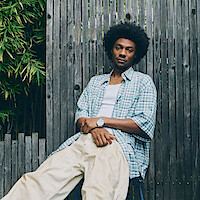 Jonathan Daviss sitting against a wooden fence outdoors, wearing a light plaid shirt, white undershirt, and cream pants, with green bamboo plants visible beside him. Relaxed expression in a natural setting.