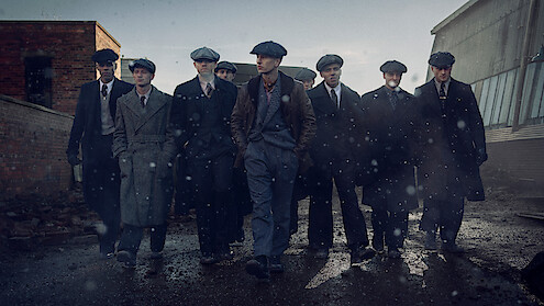 Eight men in vintage suits and flat caps walk together on a wet, gritty street in an industrial urban area, with old brick and metal buildings in the background under a gray, overcast sky.
