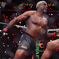 Mixed martial artist with blond hair fighting in an octagon cage during a nighttime event, wearing black shorts and red gloves, surrounded by bright arena lights and audience.