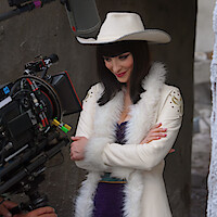 Woman in a white cowboy hat and fur-trimmed coat stands with arms crossed, smiling on a film set, as a large camera records her in an outdoor, urban environment.