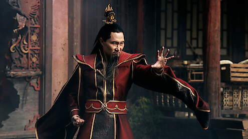 A man in an ornate red robe and crown poses with an outstretched hand in a dramatic stance, standing in a traditional Asian architectural setting with wooden pillars and stone accents in the background.