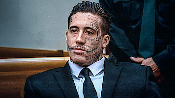 Man with extensive facial tattoos wearing a suit, seated in a courtroom setting with wooden benches and another person in uniform visible in the background.