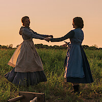 Two women in period dresses joyfully hold hands and spin in a grassy field at sunset, with warm sunlight casting a golden glow on the rural landscape.