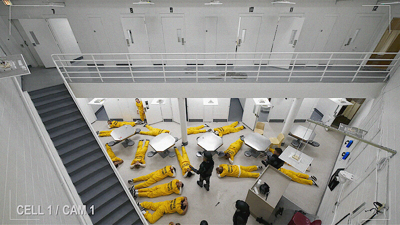 Prison common area with multiple inmates in yellow uniforms lying on the floor, viewed from above. White walls, doors, tables, and stairs in a secure facility setting. Surveillance camera footage.