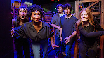 Five young adults in casual clothing look excited and curious in a colorful, dimly lit room with wooden crates and barrels, suggesting an escape room or adventure game environment.