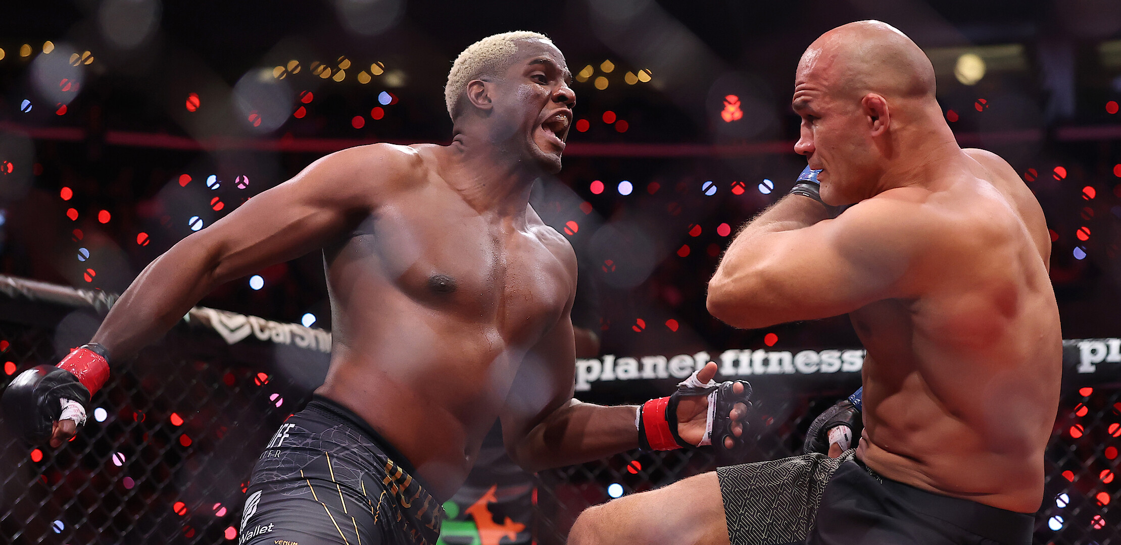 Two male MMA fighters compete intensely inside a caged octagon during a professional match, with one throwing a punch and the other defending himself. Arena lights and a crowd are visible in the background.
