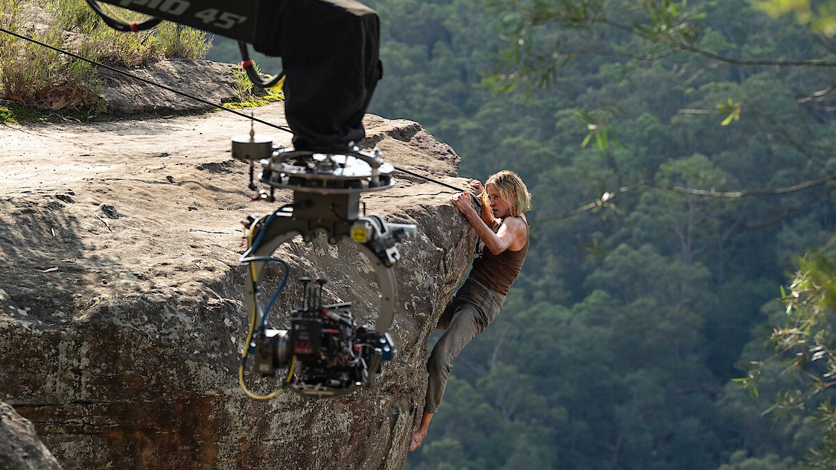 Actor climbing a rocky cliff edge in a forested area, filmed up close by a large movie camera on a crane, during an outdoor film production.