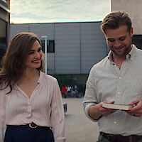 Rachel Weisz and Leo Woodall walk across campus. Woodall reads the back of a book, as both smile.