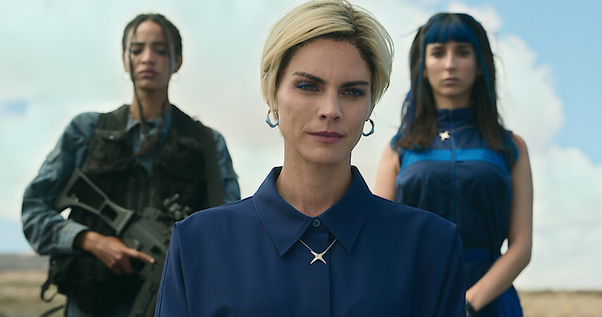 A woman stands facing the camera while to others stand behind her — one of whom is holding a rifle.