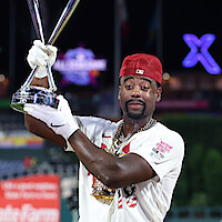 Baseball player in uniform and red cap holding trophy on field at night, stadium lights and blurred crowd in background, celebrating victory.