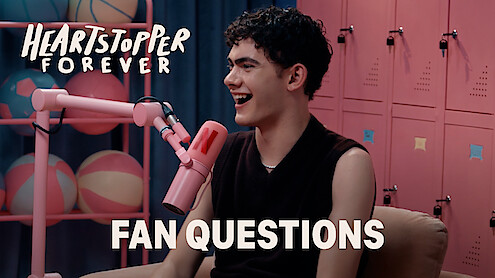 Joe Locke smiles and talks into a pink microphone in a colorful room with lockers and pastel basketballs, with "Heartstopper Forever" and "Fan Questions" text on screen, suggesting a fan Q&A interview setting.