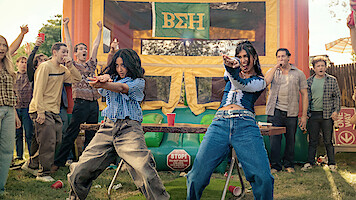 Two women dance energetically at an outdoor party with a crowd of cheering people, in front of a colorful bouncy house decorated with Greek letters, on a sunny day.
