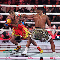 Jake Paul and Anthony Joshua fight in a brightly lit boxing ring, surrounded by ropes and an audience, with camera crews and sponsors visible in the background.
