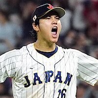 Shohei Ohtani celebrating on the field, arms raised and mouth open in excitement, wearing a white pinstripe jersey and glove, with a blurred crowd in the stadium background.