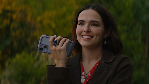 A woman smiling outdoors, holding a smartphone in her hand, wearing a brown jacket and red floral blouse, with green blurred bushes and trees in the background on a sunny day.