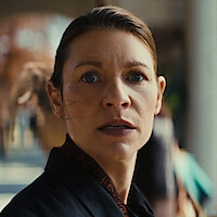 A woman with blue eyes and tied-back hair looks surprised or concerned in an outdoor public setting, with blurred people and buildings in the background.