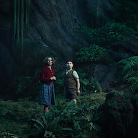  Two children stand in a lush, green forest cave with ferns and moss-covered rocks, looking up at something out of view. The scene is dimly lit, evoking a mysterious or magical atmosphere.