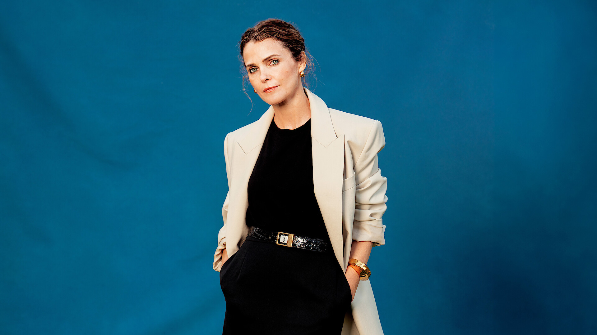 Woman posing against a solid blue background, wearing a light-colored blazer over a black outfit with hands in her pockets, accessorized with a belt and bracelet, looking relaxed and confident in a studio setting.