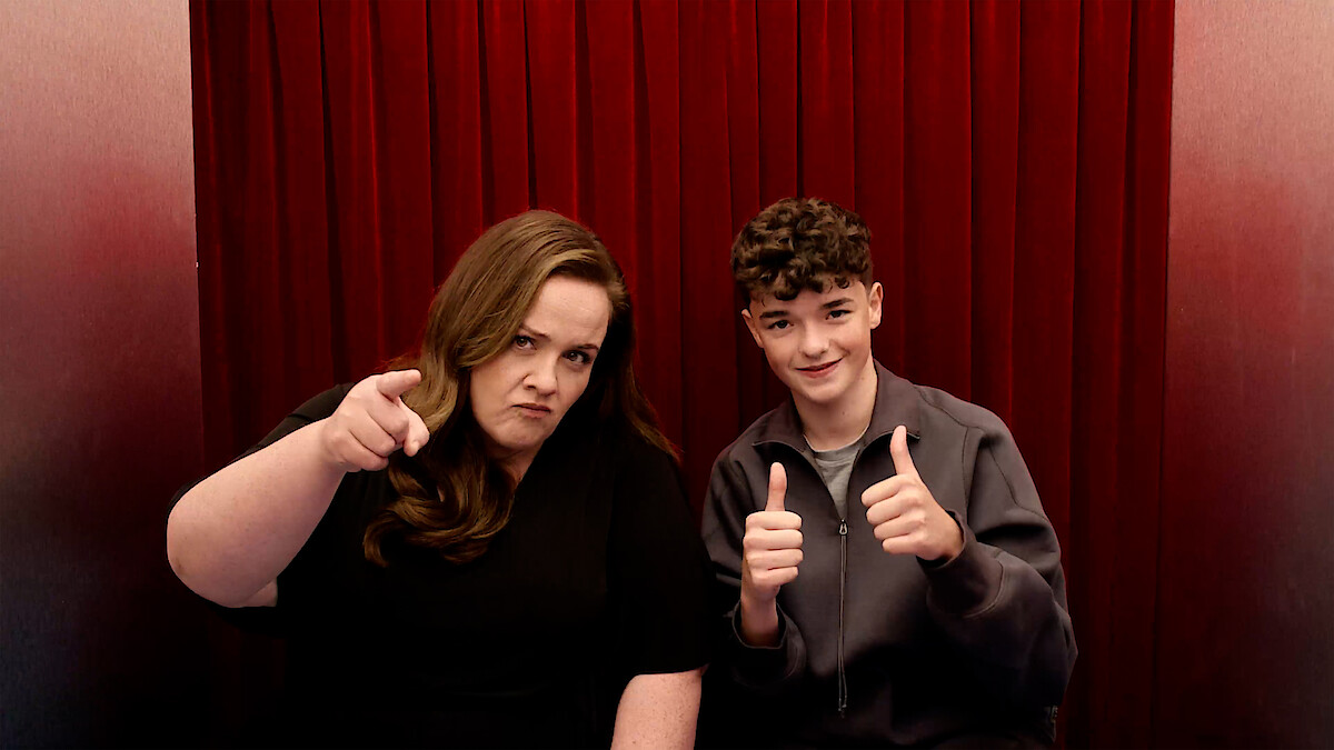 Two people sitting in front of red curtains, one woman pointing at the camera with a serious face and a teenage boy giving two thumbs up, both indoors, creating a playful and contrasting mood.