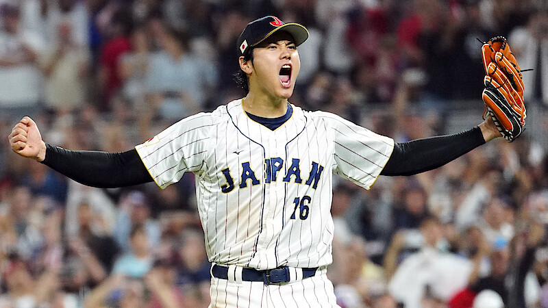 Shohei Ohtani celebrating on the field, arms raised and mouth open in excitement, wearing a white pinstripe jersey and glove, with a blurred crowd in the stadium background.