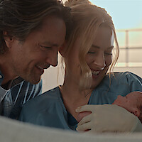 A smiling man and woman in medical scrubs hold a newborn baby together in a brightly lit room, expressing joy and tenderness after childbirth.