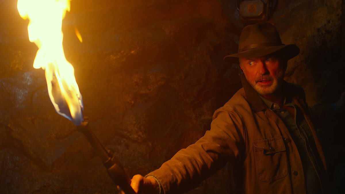 Man wearing a fedora and brown jacket holds a flaming torch in a dark, rocky cave, casting dramatic light on his determined face and the stone walls behind him.