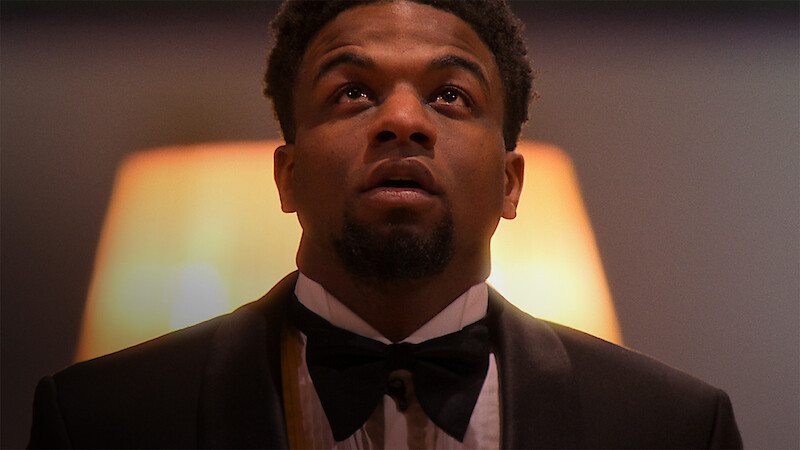 A man in a tuxedo and bowtie looks upward in a warmly lit indoor setting, appearing thoughtful or contemplative. The background is softly blurred, enhancing the formal and introspective mood of the scene.