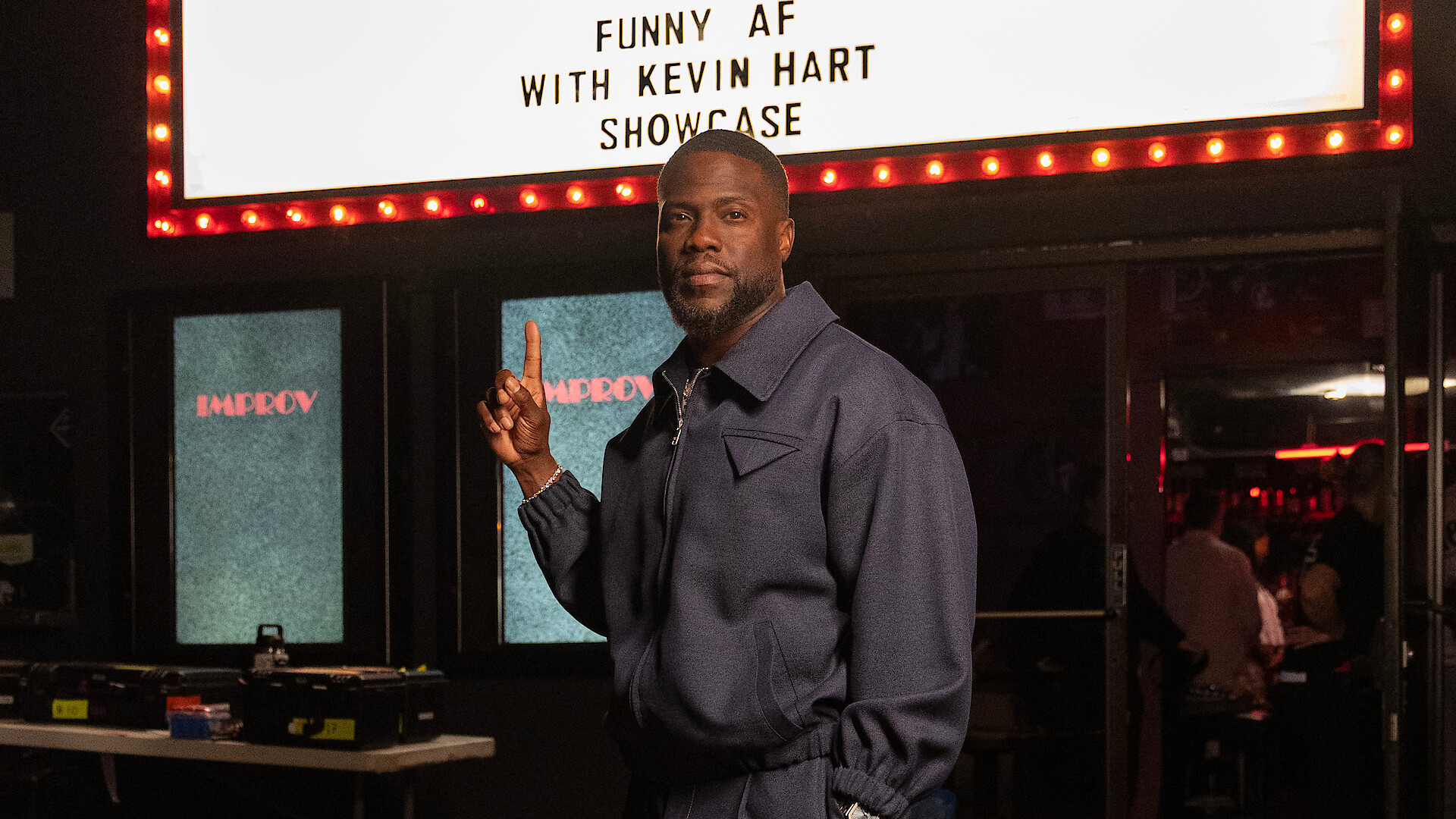 Kevin Hart in a blue jacket stands inside a comedy club, pointing upward at a lit marquee sign that reads “Funny AF with Kevin Hart Showcase,” with tables, equipment, and an audience in the background.