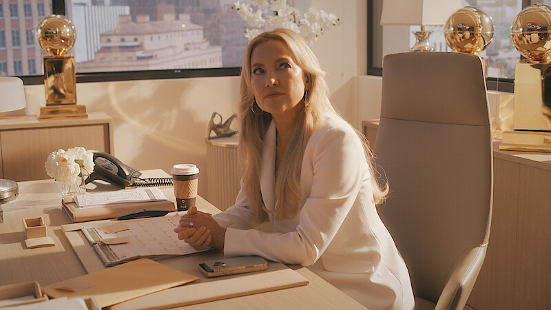 Woman with long blonde hair in a white suit sits at a modern office desk with paperwork, coffee cup, phone, and golden trophies, with cityscape visible through large windows in the background.