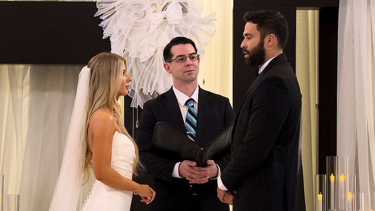 Ashley Carpenter and Alex Henderson stand facing each other in a formal indoor wedding ceremony with an officiant between them, surrounded by white drapery and candles.