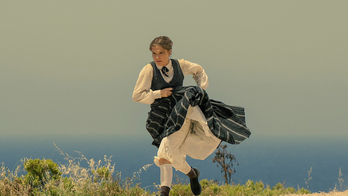 Woman in period clothing running outdoors on a grassy hill with the ocean and clear sky in the background.