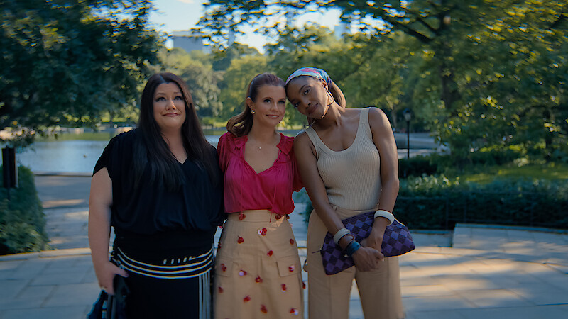 Three women standing close together outdoors on a sunny day in a lush park, smiling and posing for a photo with trees, greenery, and a body of water in the blurred background.