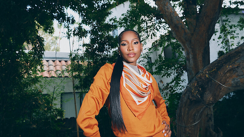 Person standing outdoors in a garden wearing an orange top and patterned scarf, posing confidently with hand on hip, large tree and house with tiled roof in the background, surrounded by greenery.