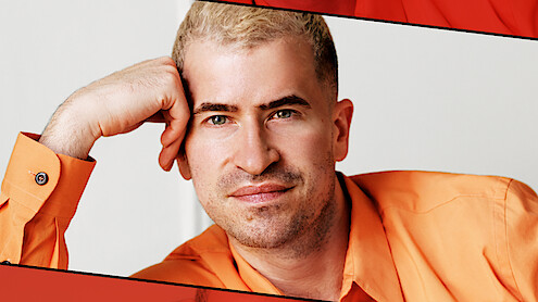 Evan Ross Katz with short blond hair in an orange shirt, resting his head on his hand, sitting indoors against a plain light background with red graphic borders.