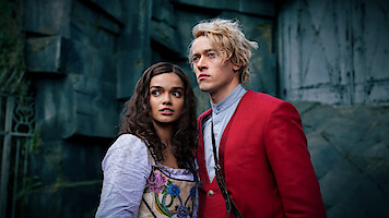 Two young adults in period costumes, a woman in a white dress with floral accents and a man in a red jacket, stand close together looking ahead, with a stone wall or ruins as the background, in a dramatic, moody setting.