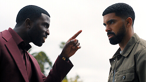 Two men outdoors, one in a maroon suit angrily pointing a finger at another man in a green jacket, who looks serious; trees and cloudy sky in the background.