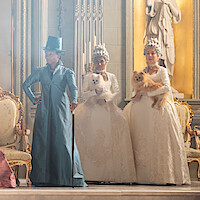 Three people in ornate period costumes stand in a lavish, gold-trimmed room. Two women in white dresses with elaborate wigs hold small fluffy dogs; another figure in blue stands beside them.