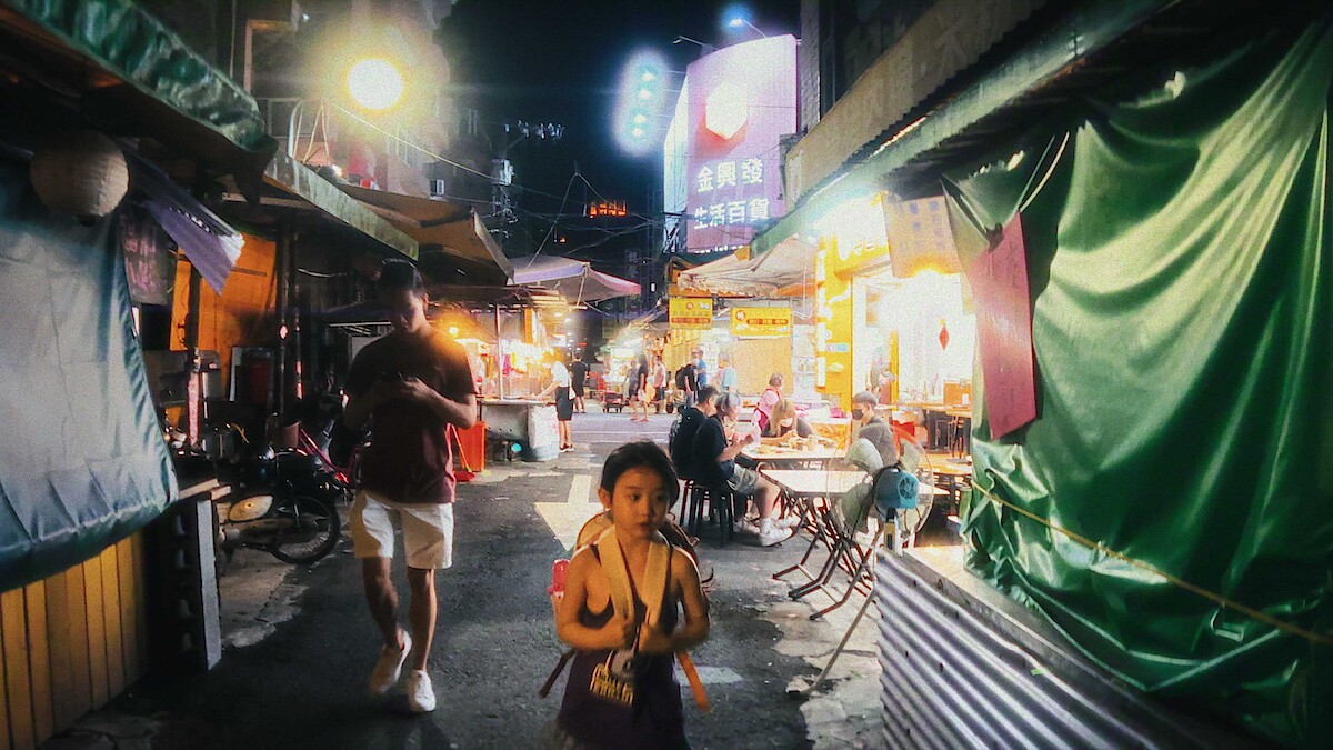 Child and adult walk through a bustling, neon-lit night market with food stalls, tables, and people dining; the scene is lively and colorful, evoking a vibrant urban atmosphere.