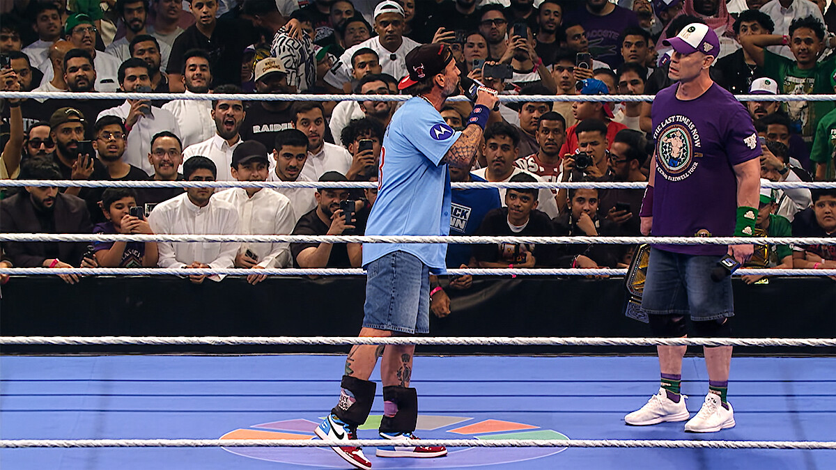 Two wrestlers stand in a ring facing each other, one holding a microphone, with a cheering audience surrounding the ring in an arena setting.