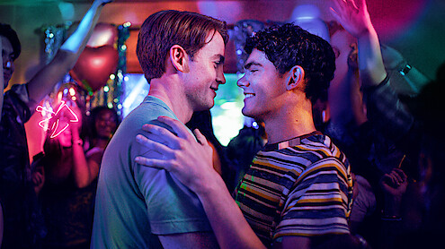 Two men smiling and dancing closely together at a vibrant, colorful party with people celebrating, arms raised, and festive decorations in the background under neon and stage lights.