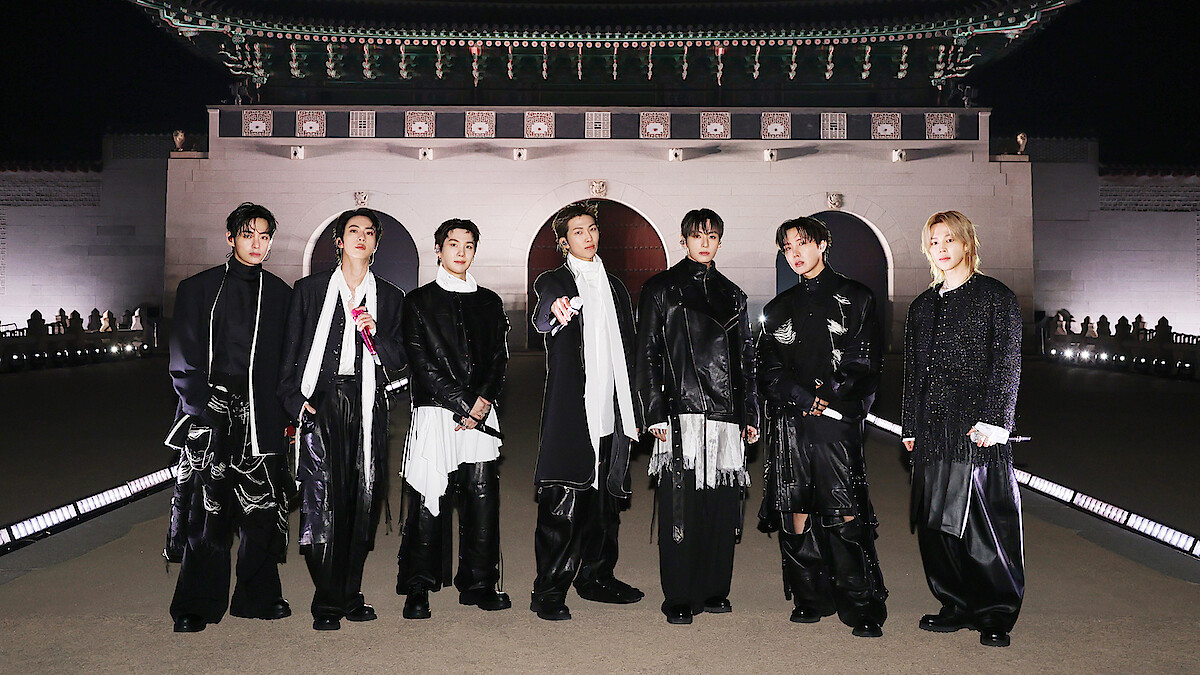 V, JIN, SUGA, RM, JUNG KOOK, J-HOPE, and JIMIN dressed in fashionable black and white outfits stand outdoors at night in front of an ornate, traditional East Asian building with arches and a large decorative roof.