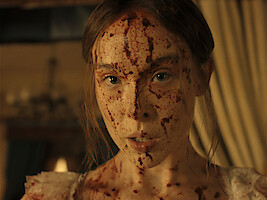 A young woman with long hair, dressed in light clothing, has her face and upper chest covered in blood, standing indoors with dim warm lighting and blurred curtains and furnishings in the background.