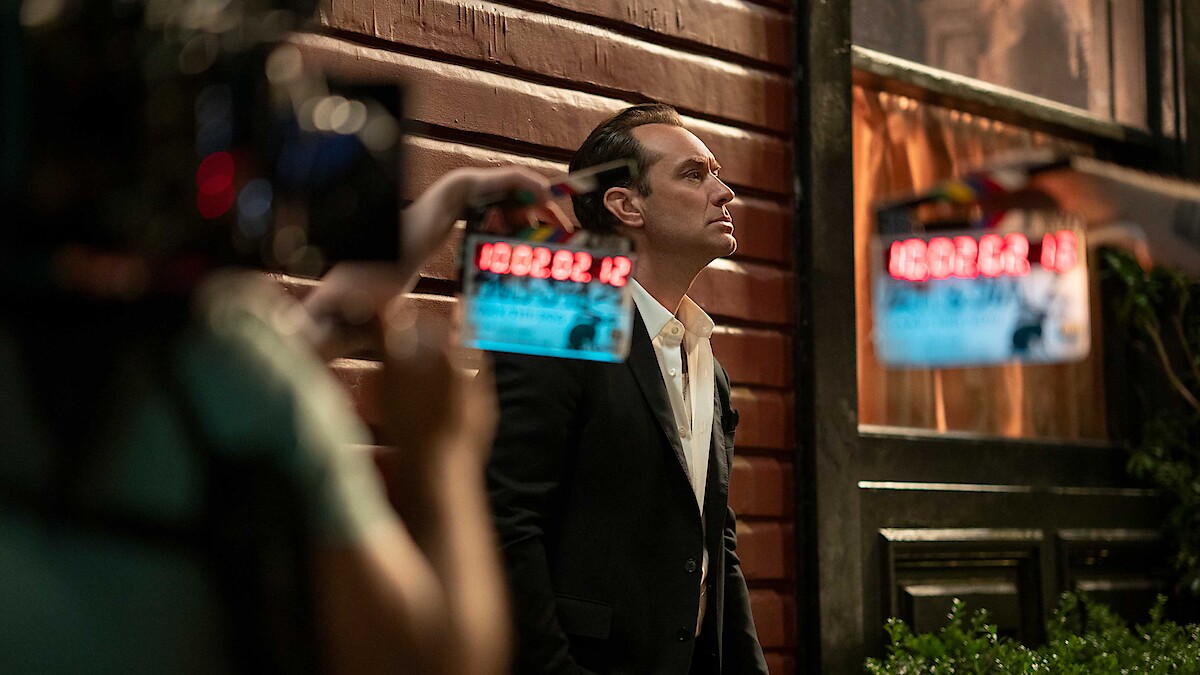 A man in a suit stands against a wooden wall at night, looking up thoughtfully. Movie cameras and crew are visible, suggesting a film set environment. Warm lighting from windows creates a cinematic atmosphere.
