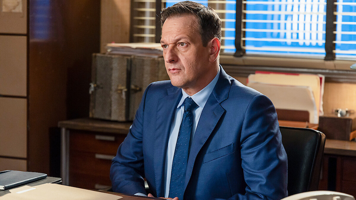 Man in a blue suit sitting at a desk in an office environment, with files, folders, and boxes on wooden furniture, blinds on the window letting in daylight behind him.