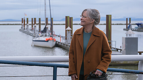 Sally Field as Tova in a brown coat standing by a railing at a marina, with sailboats docked and calm water in the background under a cloudy sky.