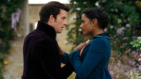 A man and woman in period clothing stand close together, gazing into each other's eyes in an outdoor garden with stone walls and blooming flowers, conveying a dramatic or romantic moment.