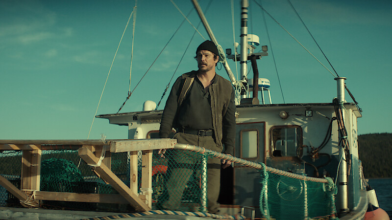 A man wearing a beanie stands on the deck of a fishing boat surrounded by nets and wooden railings, with the sea and distant land visible under a clear sky.