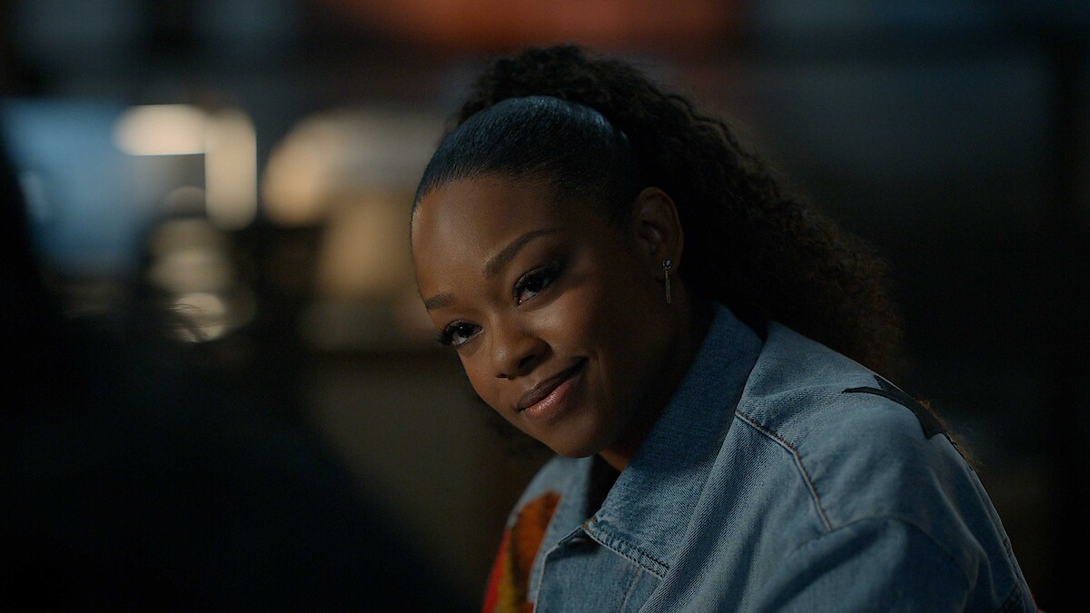 A woman with curly hair tied back is sitting indoors, wearing a denim jacket, and smiling warmly in a softly lit environment with a blurred background.